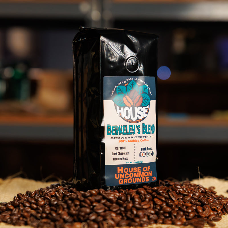 A bag of House of Uncommon Grounds Berkeley's Blend coffee stands upright on a burlap sack surrounded by loose roasted coffee beans, with a blurred bar or kitchen background.