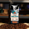 A bag of House of uncommon grounds Sunrise Espresso whole bean coffee stands upright on a burlap sack surrounded by scattered roasted coffee beans, with a blurred bar or kitchen background.