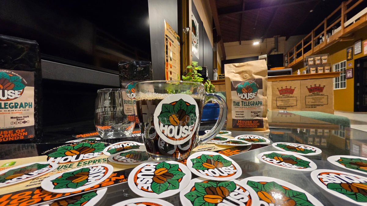 A glass mug of House coffee among House stickers, with bags of The Telegraph and the roastery floor behind